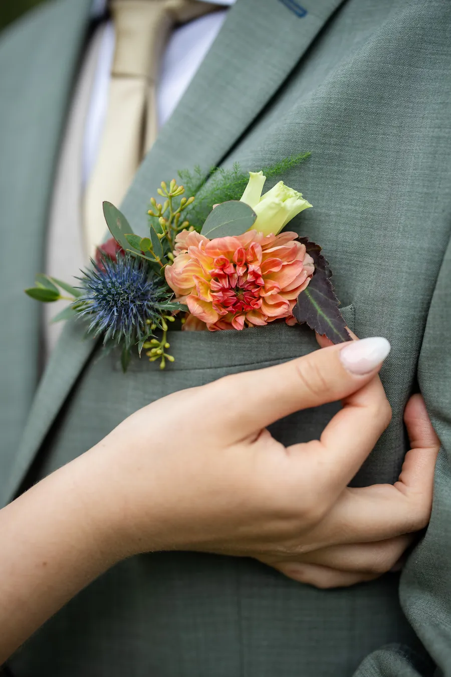 Detaljbilde av brudgommens corsage med blå tistel og oransje zinnia på salviegrønn jakke
