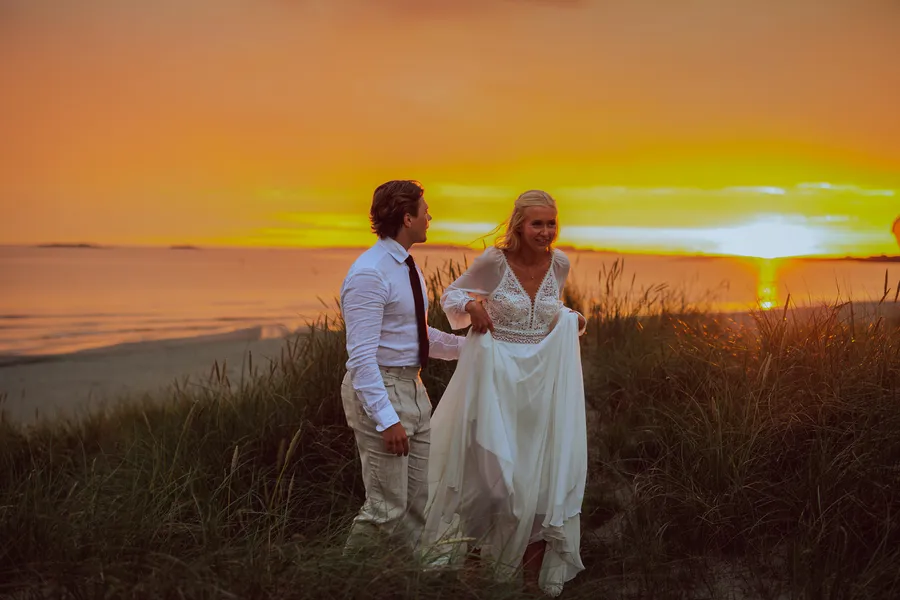 Brudepar i sanddyner med dramatisk oransje solnedgang over havet