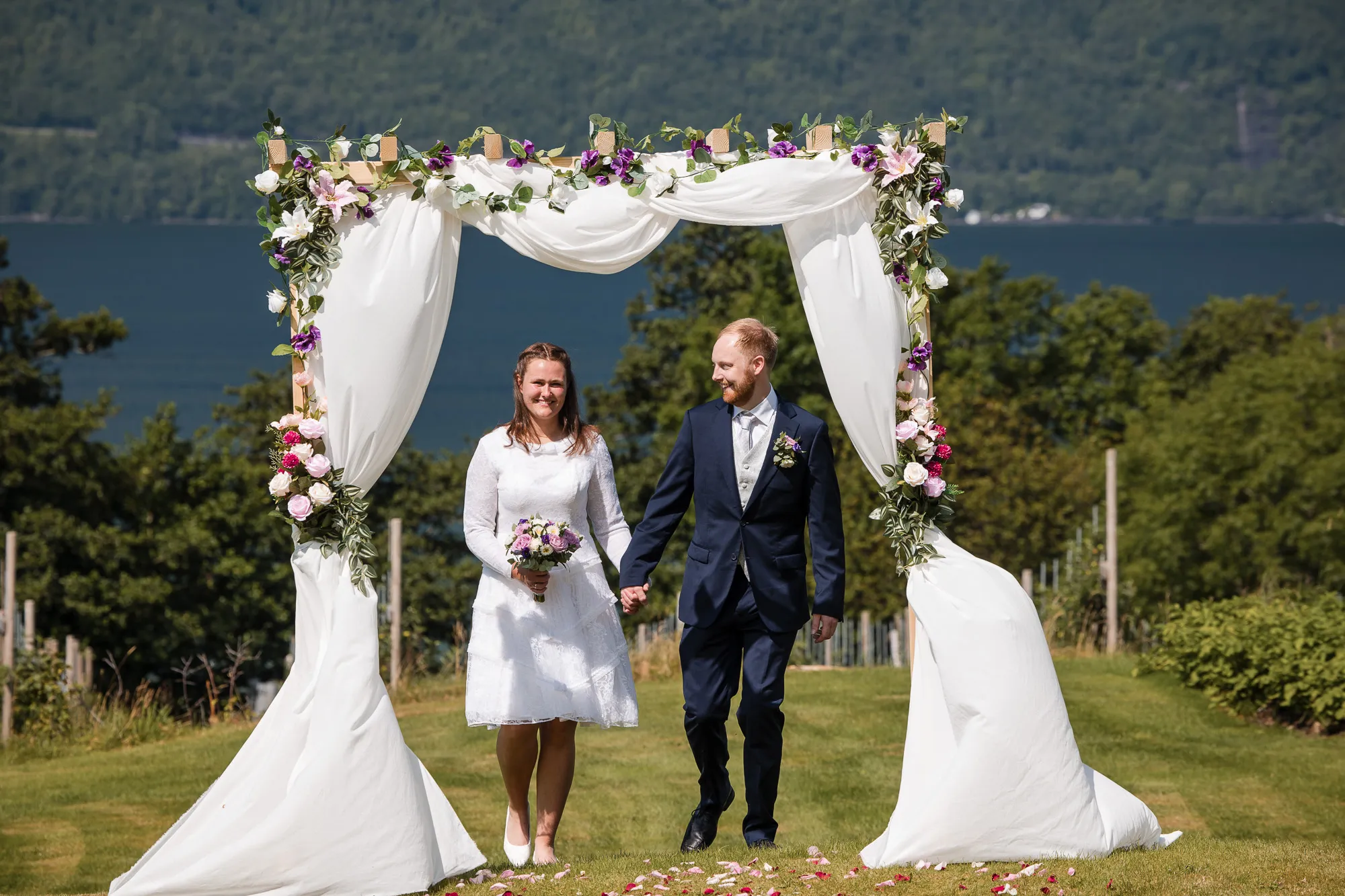 Kristine og Lars under en blomsterbue med Hardangerfjorden i bakgrunnen — bryllupsfotograf Hardanger