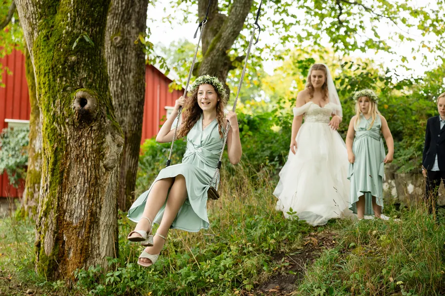 Brudepike med blomsterkrans på huske mens bruden går forbi i hagen